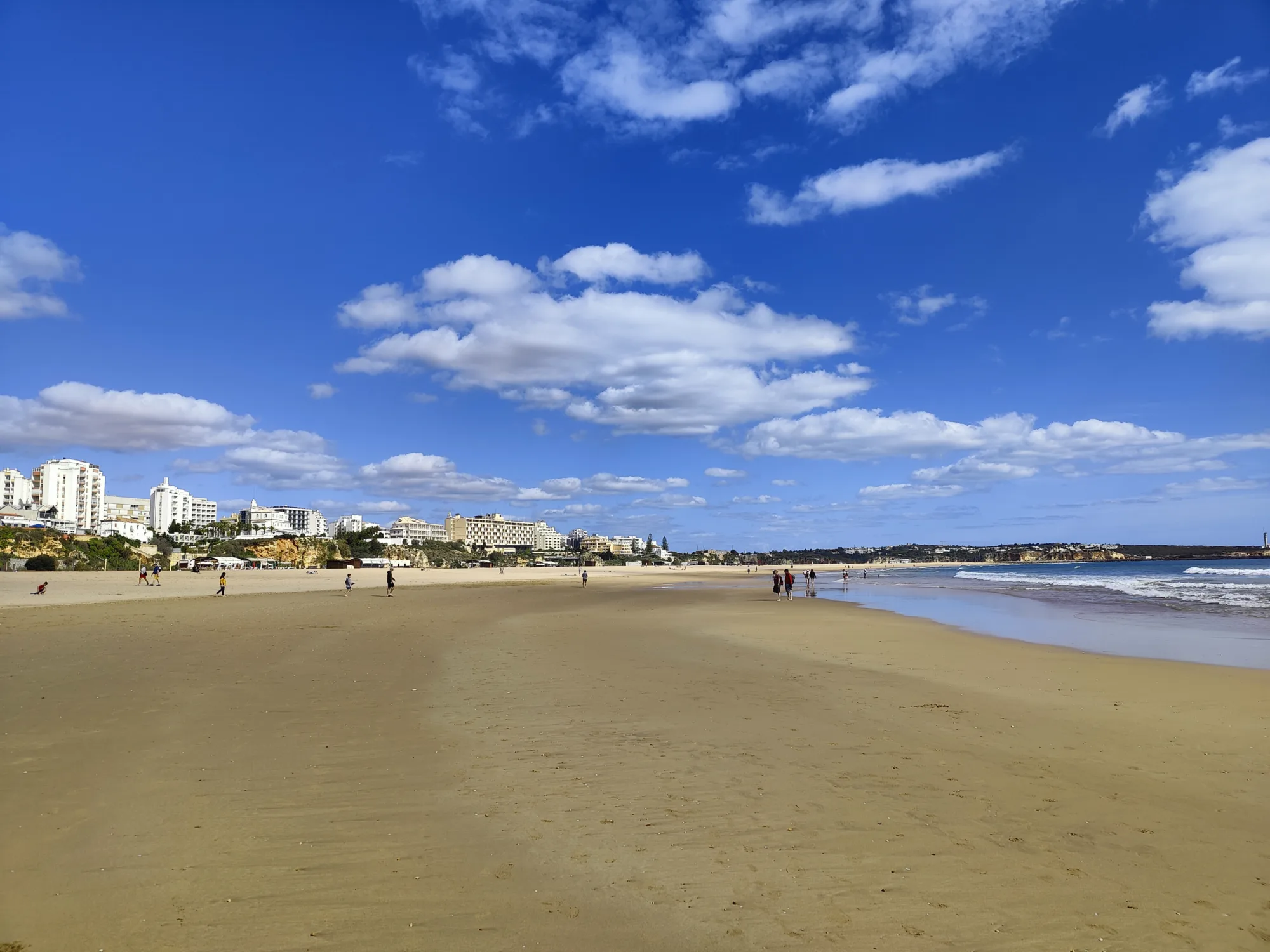 a sandy beach with people walking on it and buildings in the background